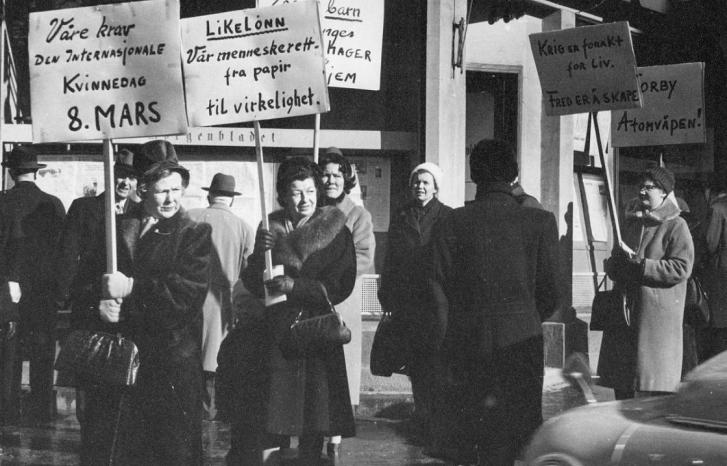 Åtte kvinner går langs fortauet med plakater på det som trolig er Norges første 8. mars markering. Parolene handler om likelønn, fred og nei til atomvåpen