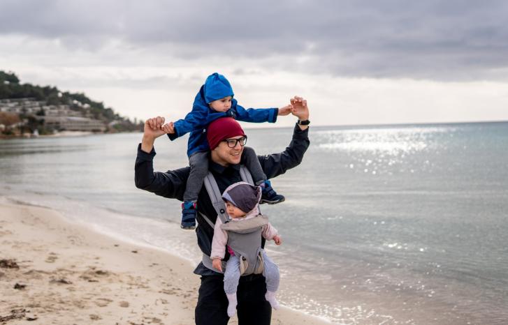 Pappa på tur på stranden med ett barn i bæresele og ett på nakken