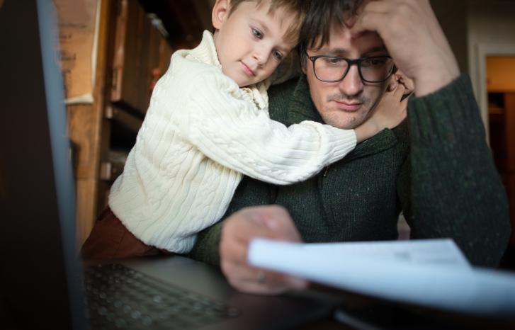 Mann sitter foran en dataskjerm og holder et ark, og ser stresset ut. Et barn holder rundt ham. 