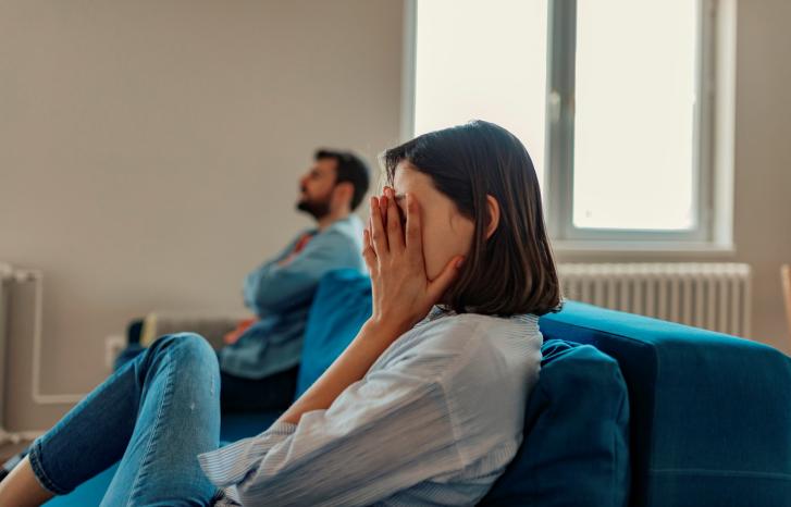 Kvinne i blå sofa med hendene foran ansiktet. Rådvill mann i bakgrunnen.