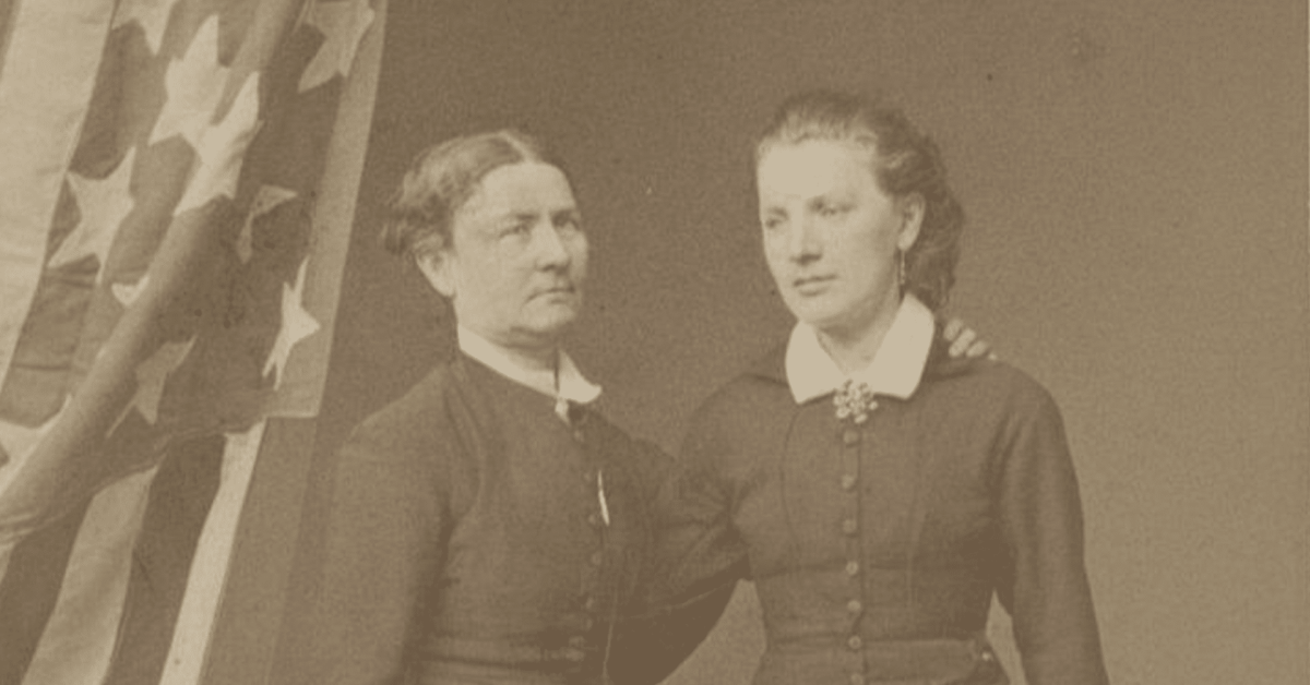 Svart-hvitt foto av Aasta Hansteen og Theodora Nilsen med et amerikansk flagg i bakgrunnen.