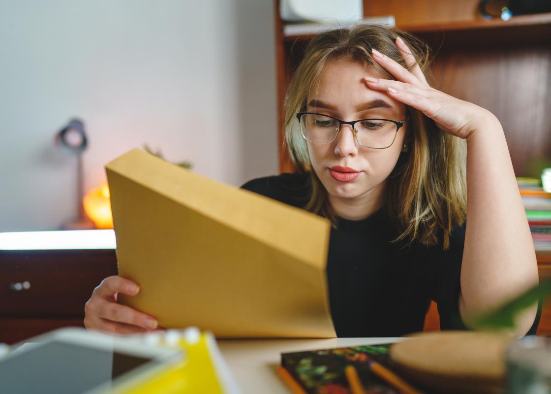 Jente eller ung kvinne med blondt og hår og briller som ser stresset ut og holder papir i den ene hånda