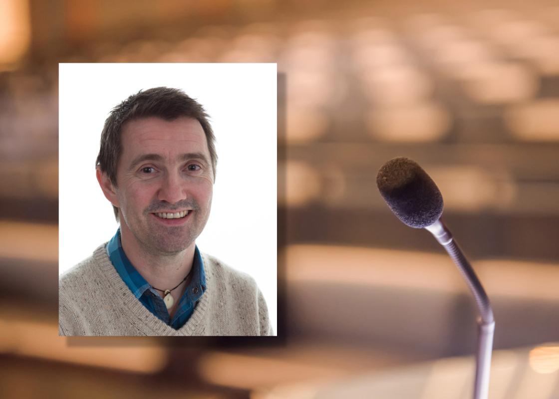 Portrettfoto av Torjer Olsen mot bakgrunn av en mikrofon og et tomt auditorium