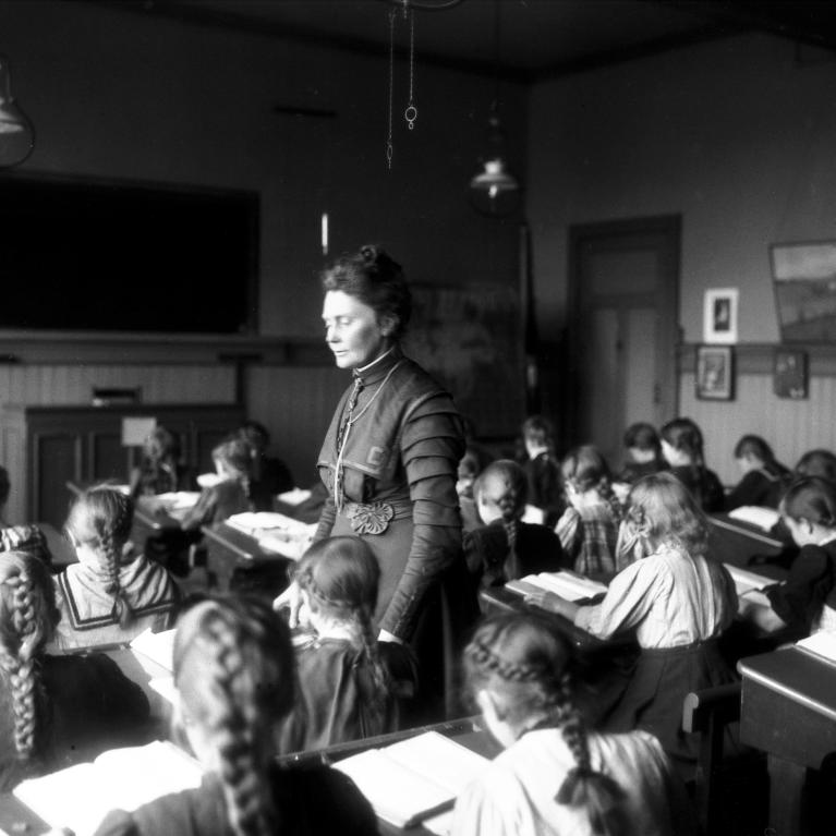 Sort-hvitt-foto av Anna Rogstad stående i klasserommet mens barna sitter bøyd over pultene sine i 1909
