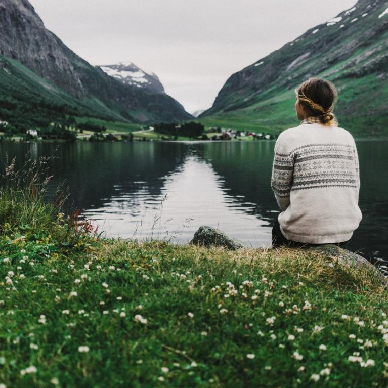 Ung kvinne sitter med ryggen til ved et vann omringet av fjell